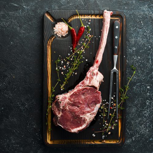 Dry aged raw tomahawk beef steak with ingredients on a black stone background. Top view. Flat Lay.